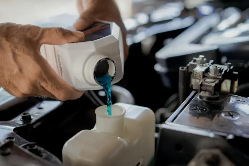 Hands pour blue coolant into a car's reservoir. This maintenance task is essential for preventing overheating, which can lead to costly car radiator replacement.